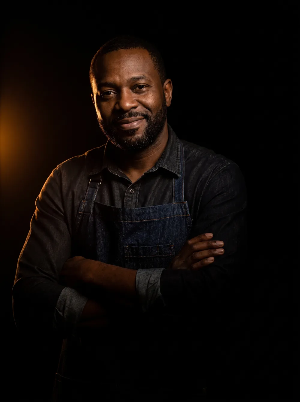 A barbershop owner in warm low light, arms crossed, fading into the dark